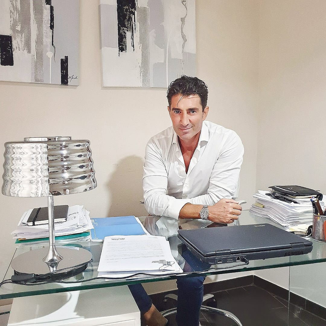 Uomo sorridente seduto alla scrivania con laptop, documenti e lampada. Ufficio con quadri.