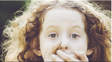 Bambina con mani sulla bocca. Linguaggio del corpo: silenzio, segreto. Capelli ricci.
